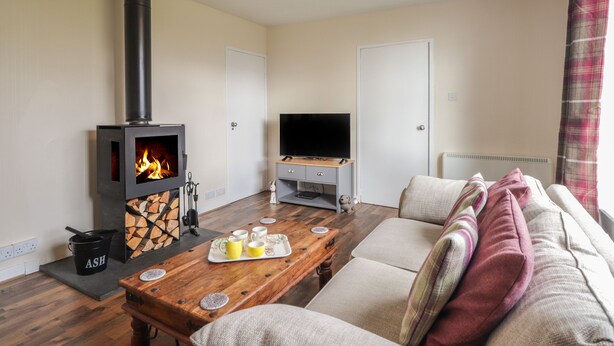 A cozy living room with a wood-burning stove, a television, a sofa, and a dining table. - Taigh Neilag (Strath)