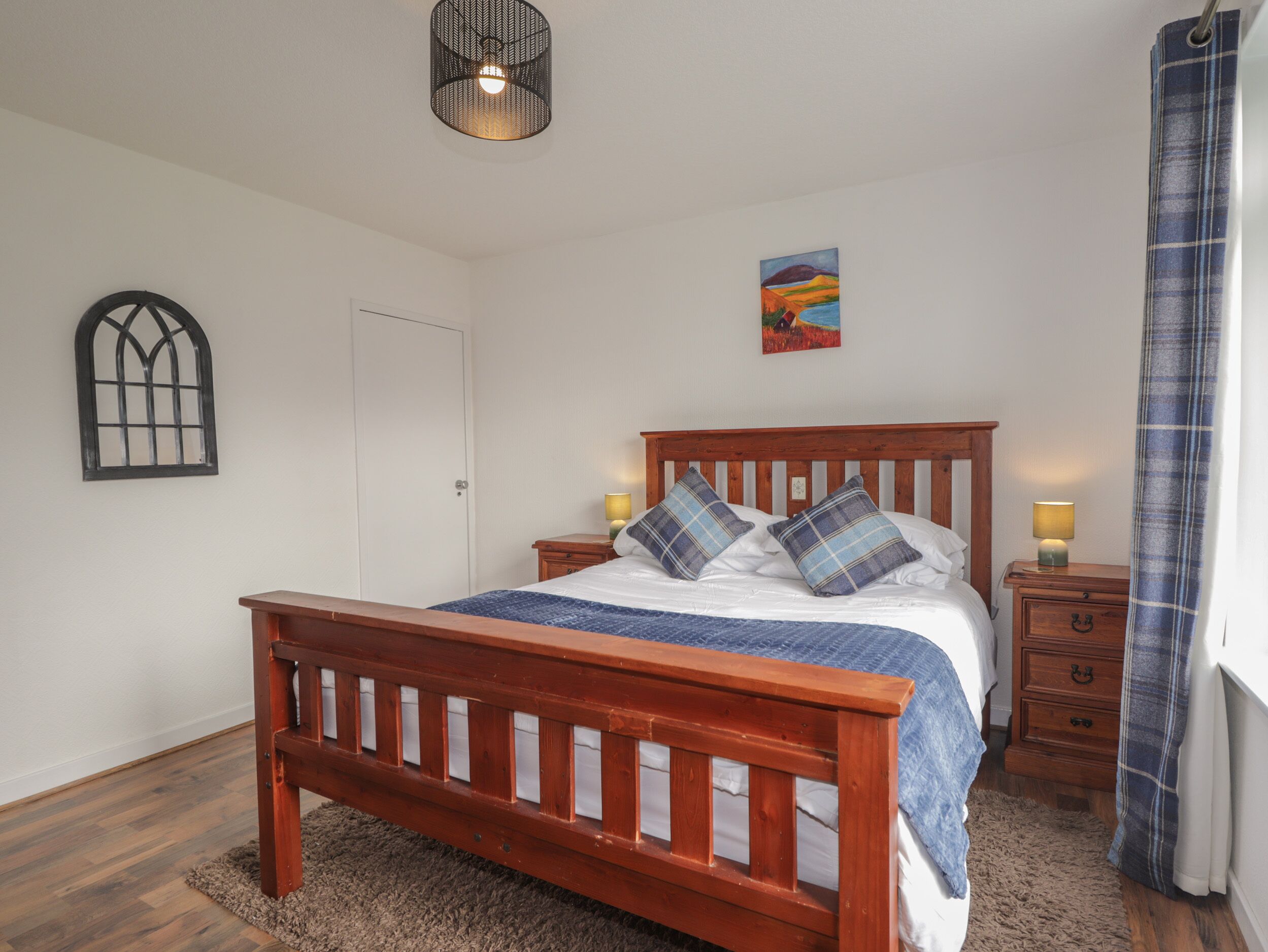 A bedroom with a wooden bed, two bedside tables, a nightstand, a window with a metal frame, a painting on the wall, and a curtain on the window.