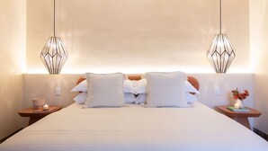 Ein ordentlich bezogenes Bett mit weißen Leinen und Kissen, zwei Nachttische mit Kerzen sowie geometrische Pendelleuchten.