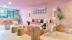 Ein modernes Wohnzimmer mit einer rosa Wand, einem Sofa, einem Couchtisch, Rattan-Hockern und einem großen Fenster mit Blick auf Bäume.