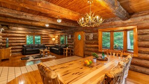 A log cabin interior with a wooden dining table, a black leather sofa, and a chandelier. - Log Cabin with Private Indoor Heated Pool-Hot Tub-firepit-Game Room-100 acres (Homer)