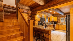 A log cabin interior with a wooden staircase, a kitchen area with wooden cabinets, a bar counter, and a refrigerator. - Log Cabin with Private Indoor Heated Pool-Hot Tub-firepit-Game Room-100 acres (Homer)