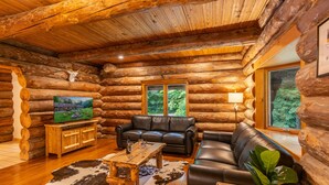 A log cabin interior with a wooden ceiling, a leather sofa, a wooden coffee table, and a television on a wooden stand. - Log Cabin with Private Indoor Heated Pool-Hot Tub-firepit-Game Room-100 acres (Homer)