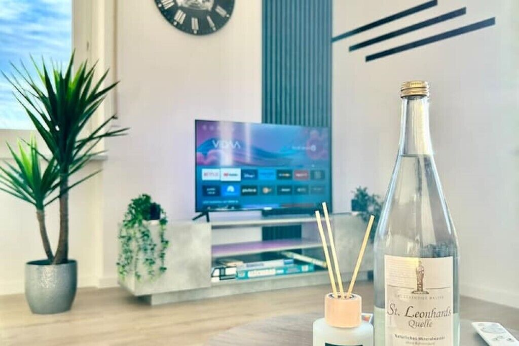 A modern living room with a television, a bottle of St. Leonhard's Quelle mineral water, and two potted plants.
