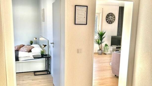 A modern bedroom with a bed, bedside table, and lamp. A hallway leads to a living area with a TV, sofa, and plants. - Diamond Apartments: Dreiländereck Nahe Flughafen (Rheinfelden (Baden))