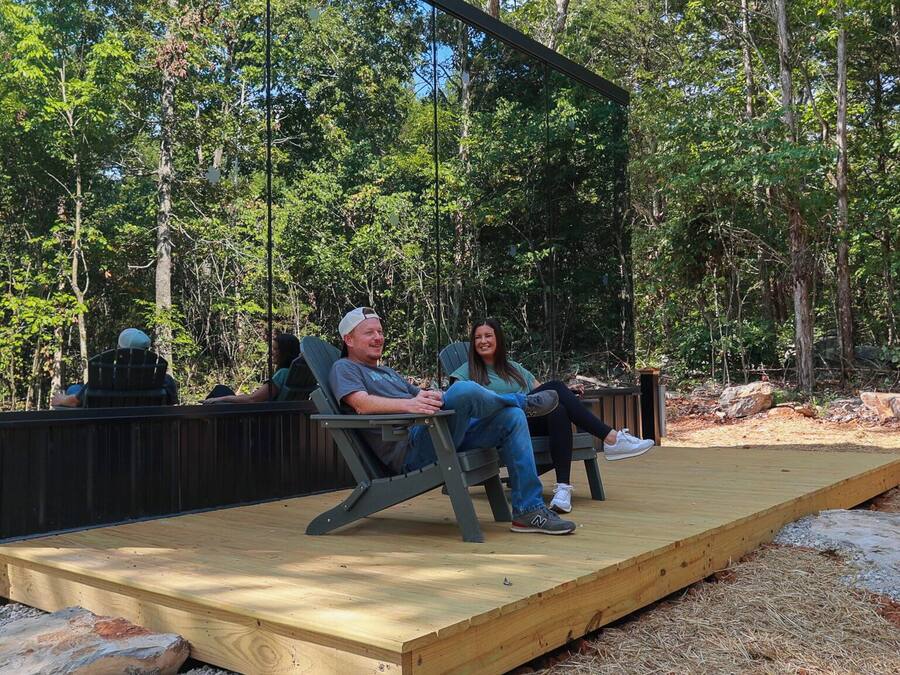 A wooden deck with two people seated on chairs, surrounded by a forested area.