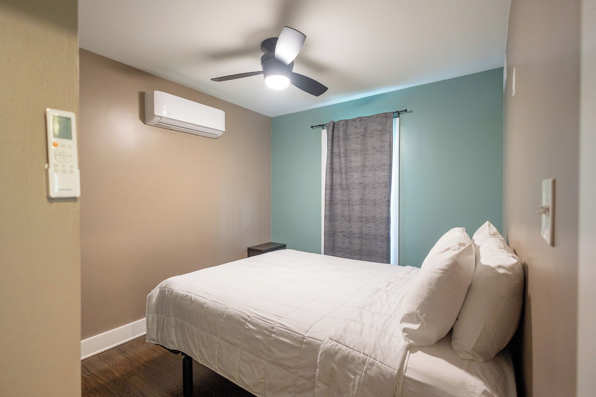 Quarto com cama, ar condicionado, ventilador de teto, janela com cortinas e uma mesinha de cabeceira.