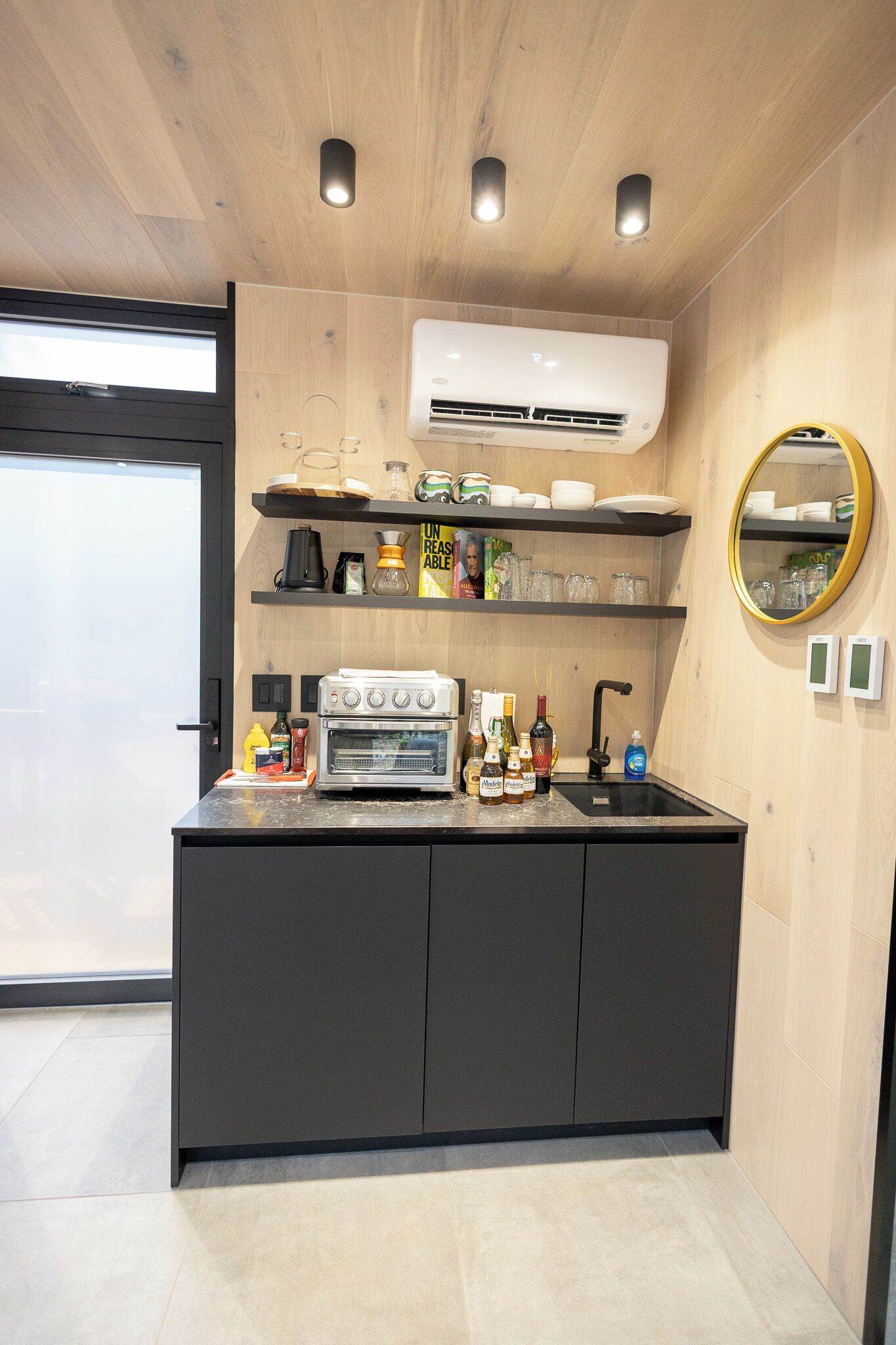 A modern kitchen with a built-in oven, a coffee machine, and a sink. There are shelves above the counter holding various kitchen items.