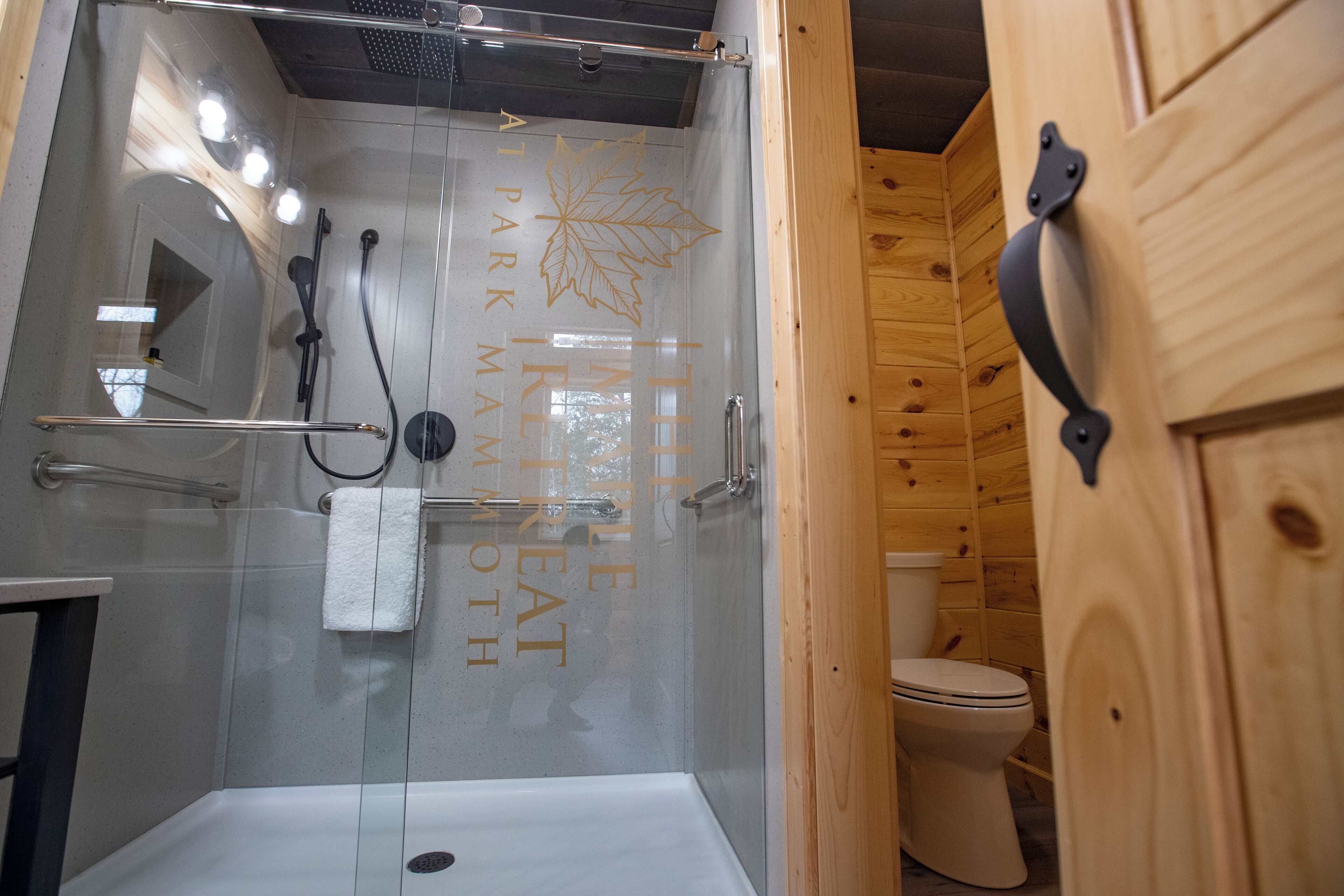 Ducha envuelta en vidrio con bañera blanca, barra para toallas y retrete en un baño estilo cabaña de madera.