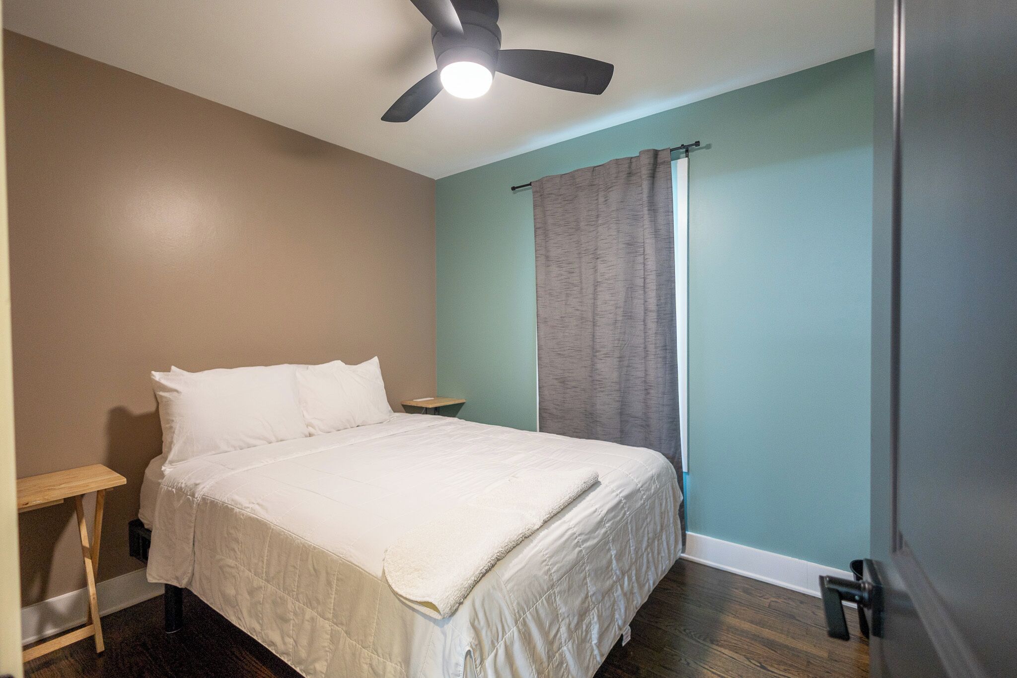 Quarto com cama, ventilador de teto, mesa de cabeceira e janela com cortinas.