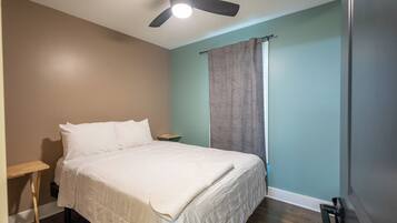 Quarto com cama, ventilador de teto, mesa de cabeceira e janela com cortinas.