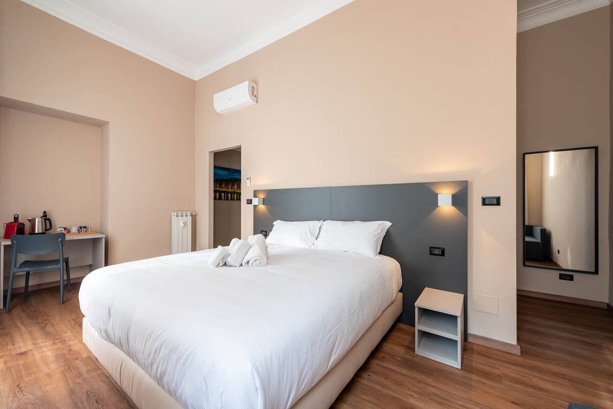 A modern hotel room with a large bed, a desk with a chair, a small table, and a mirror.
