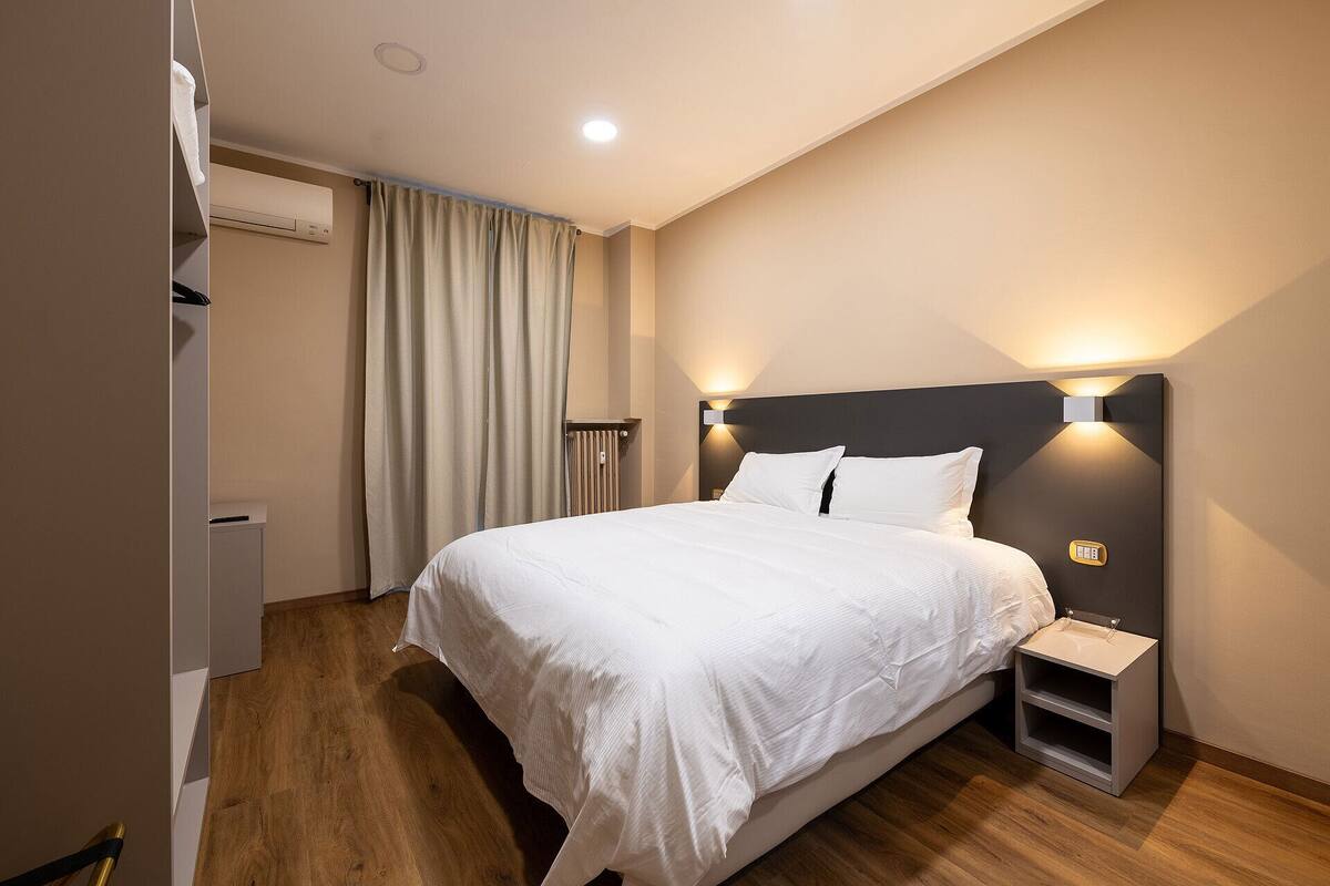 A hotel room with a large bed, a bedside table, a wall-mounted light, and a radiator.