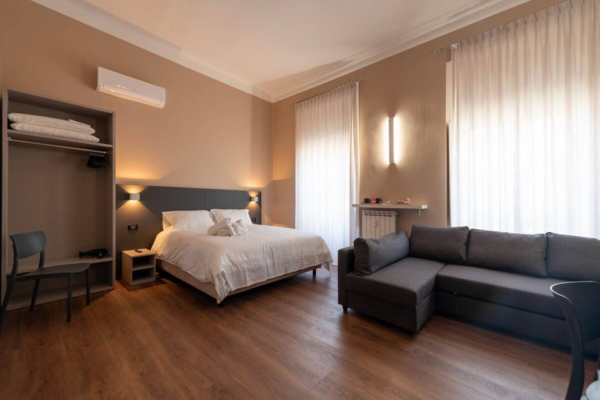 A hotel room with a bed, a sofa, a wardrobe, a desk, and a window with curtains.