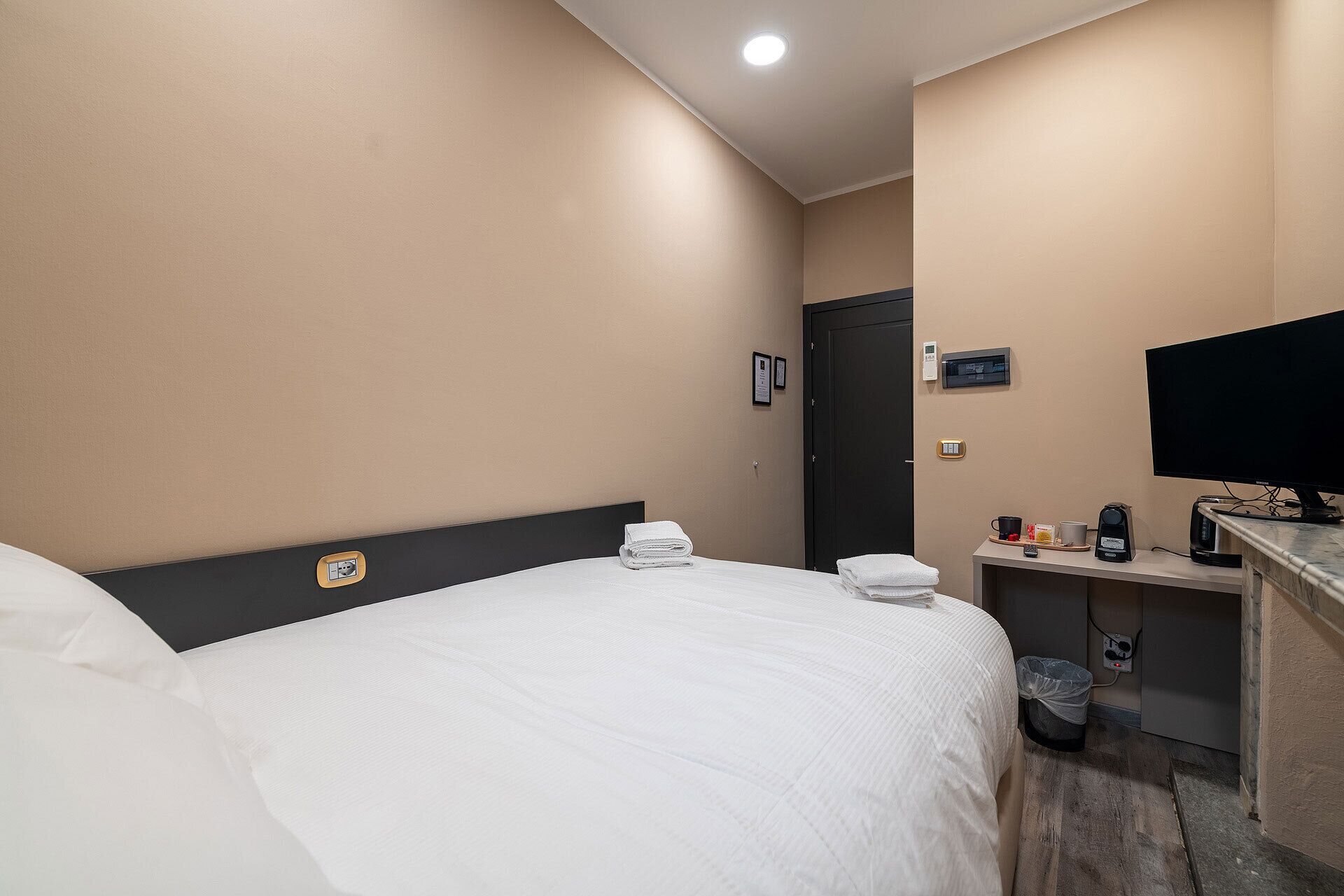 A hotel room with a bed, a desk with a coffee maker, a trash bin, and a TV on the wall.