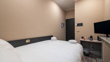 A hotel room with a bed, a desk with a coffee maker, a trash bin, and a TV on the wall.