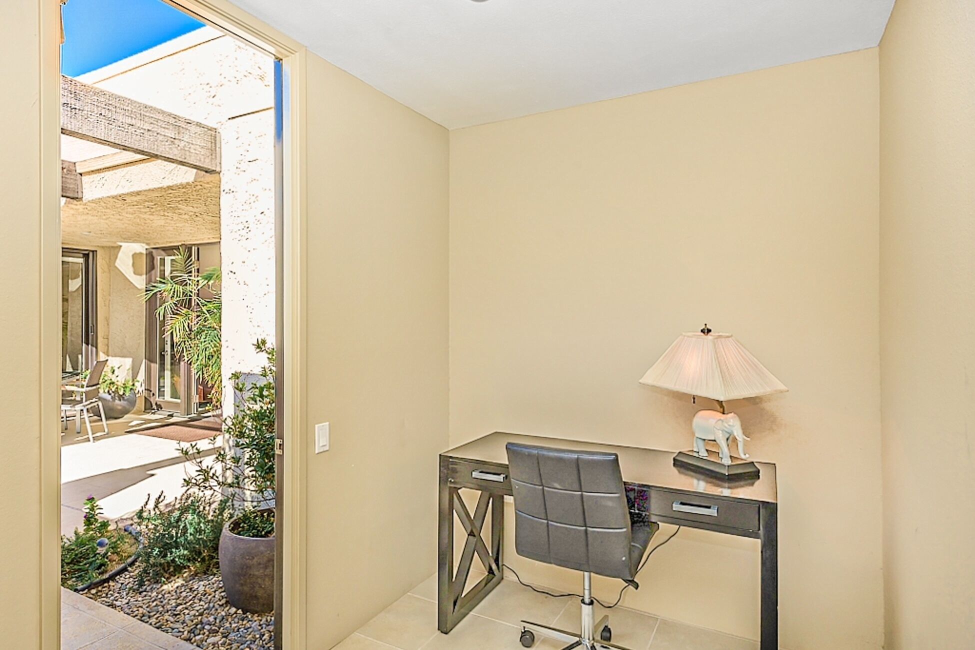 A small office space with a desk, chair, and lamp. There is a view of a patio with plants and a building outside.