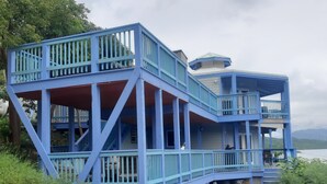 Un edificio azul de varios niveles con balcones, un paseo cubierto y vista al agua.