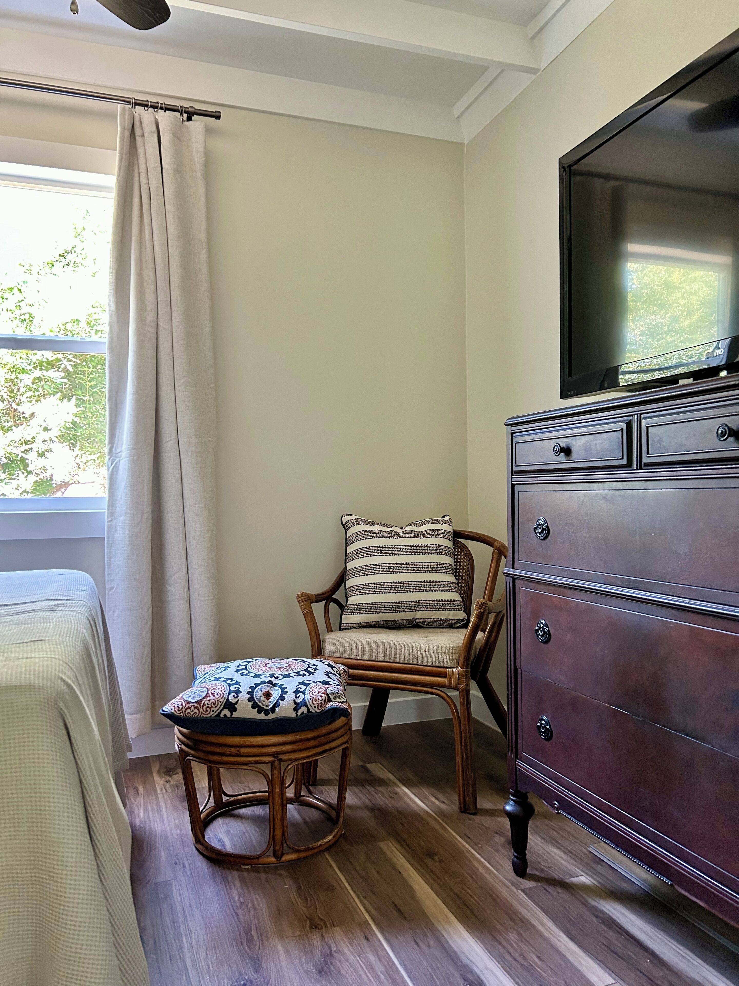 A room with a wooden floor, a wicker chair with a patterned cushion, a wooden dresser, and a window with a curtain.