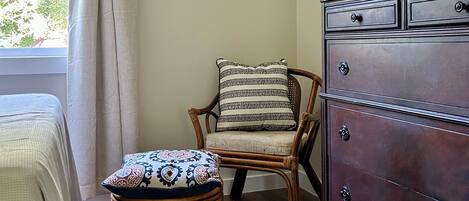 A room with a wooden floor, a wicker chair with a patterned cushion, a wooden dresser, and a window with a curtain.