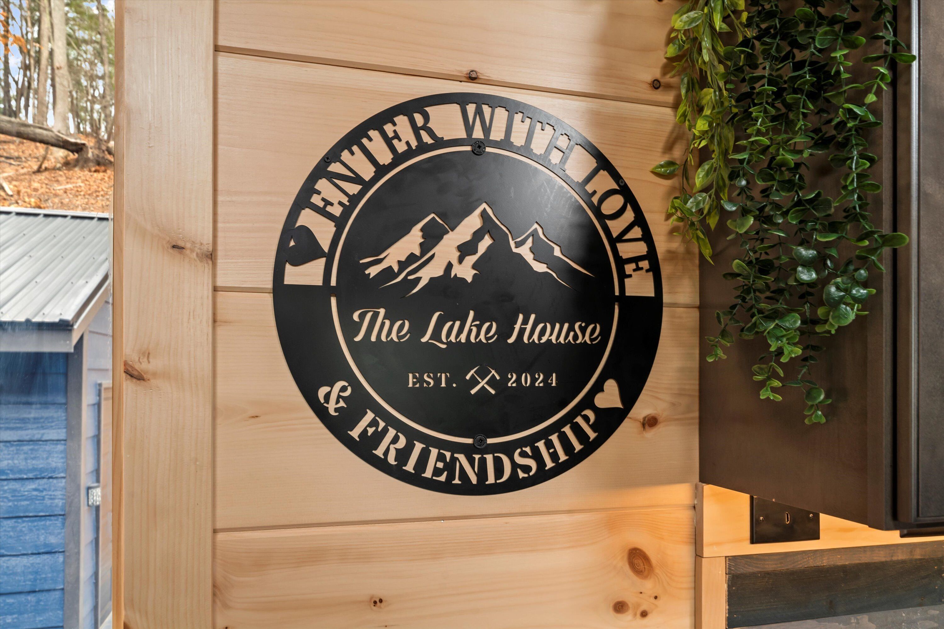 Ein hölzernes Schild mit einem kreisförmigen Design, das eine Bergzeichnung und den Text 'The Lake House & Friendship' sowie das Gründungsjahr 2024 zeigt.