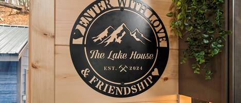 Ein hölzernes Schild mit einem kreisförmigen Design, das eine Bergzeichnung und den Text 'The Lake House & Friendship' sowie das Gründungsjahr 2024 zeigt.