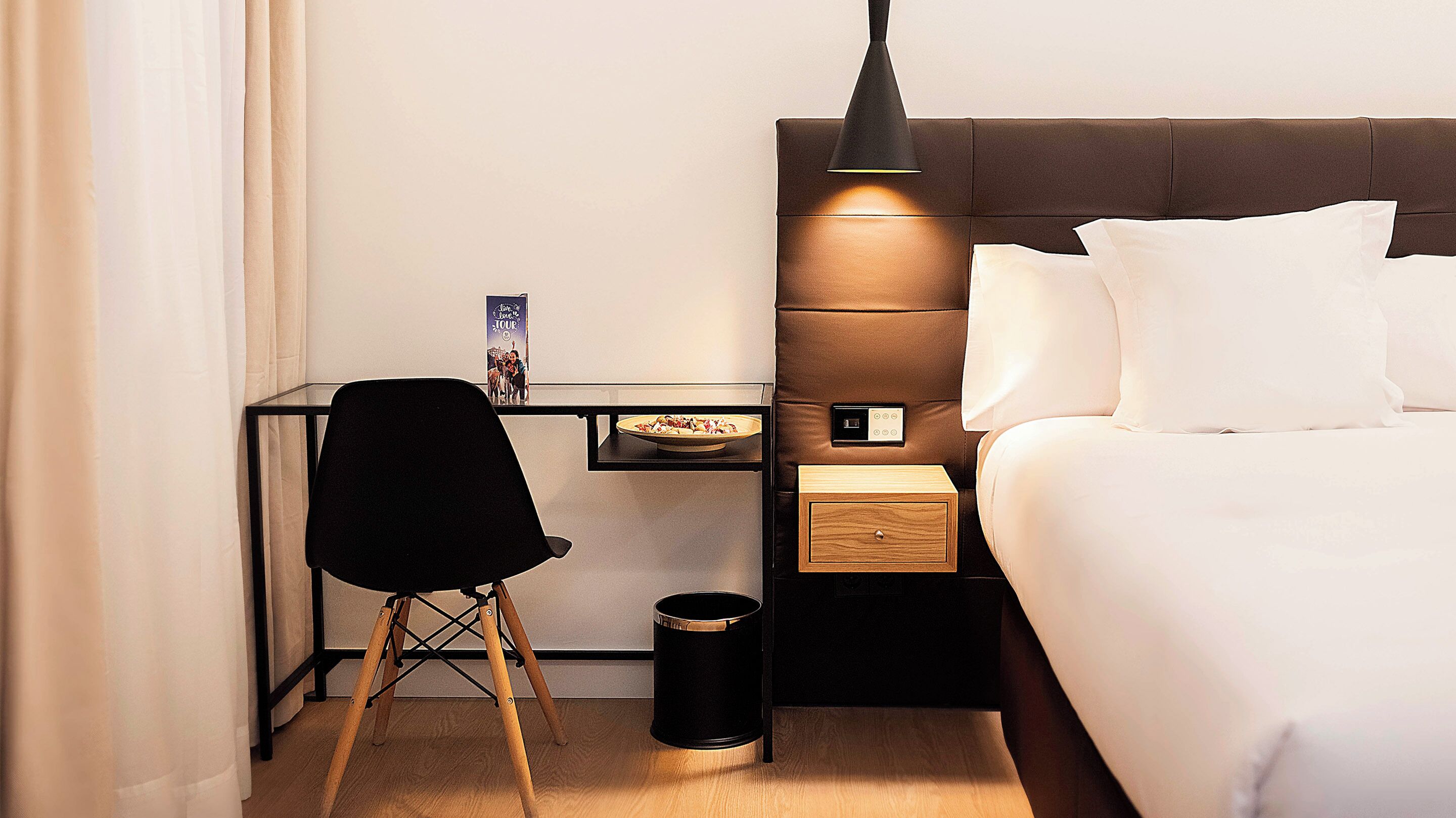 A hotel room with a bed, a desk with a chair, a small table with a plate of food, a trash can, and a wall-mounted light fixture.