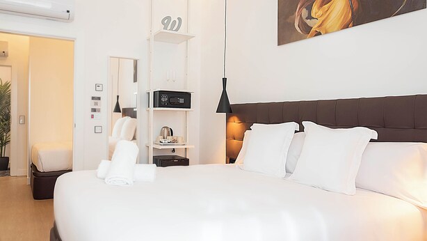 Una habitación de hotel moderna con una cama grande, una mesita de noche con una radio, un pequeño estante con objetos decorativos y un cuadro en la pared.