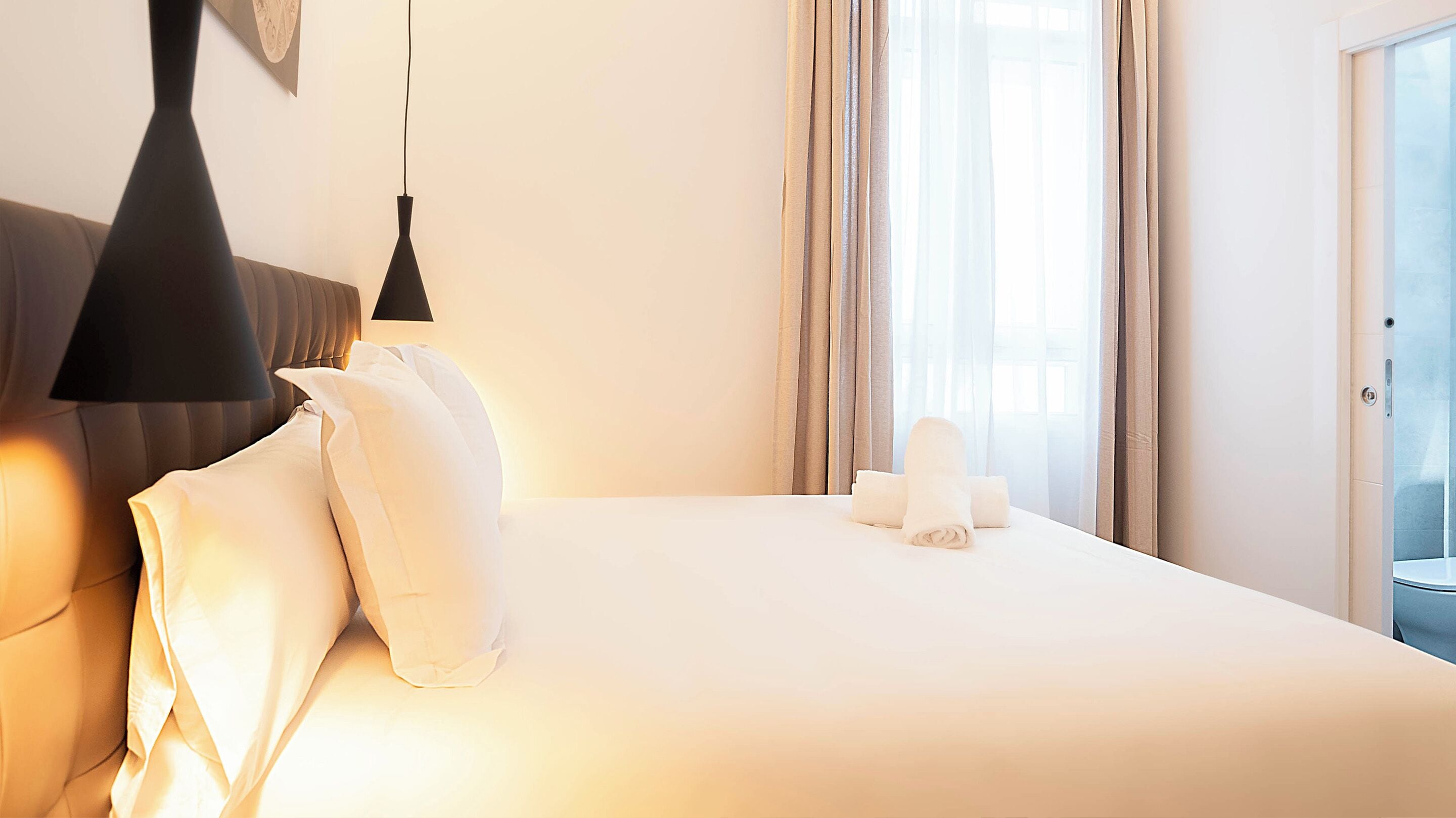 A hotel room with a neatly made bed, a hanging lamp, and a view of the outdoors through the window.