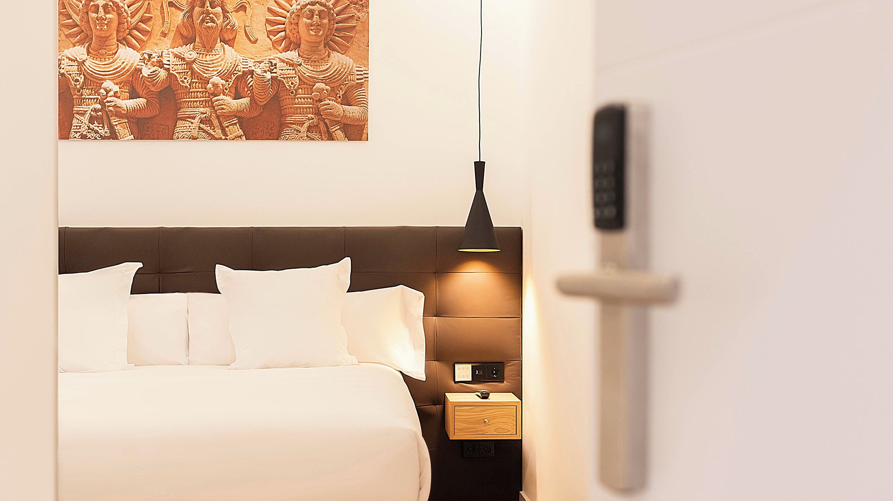 Ein modernes Hotelzimmer mit einem großen Bett, einem Nachttisch, einem an der Wand befestigten Kunstwerk und einer Pendelleuchte.
