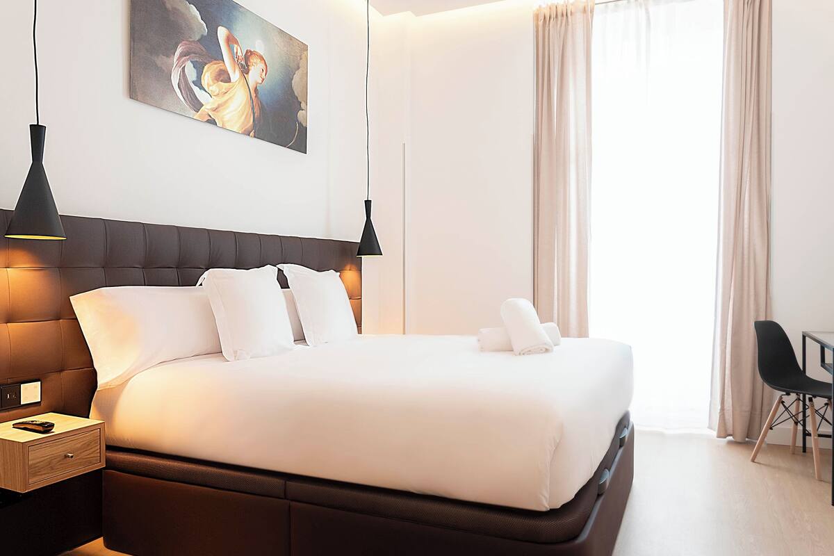 Een netjes opgemaakt bed met witte lakens, een donker hoofdbord en twee zwarte hanglampen.