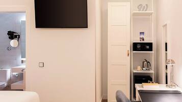 Una camera d'albergo moderna con televisione a schermo piatto fissata al muro, macchina da caffè integrata e un piccolo ripiano con oggetti personali.