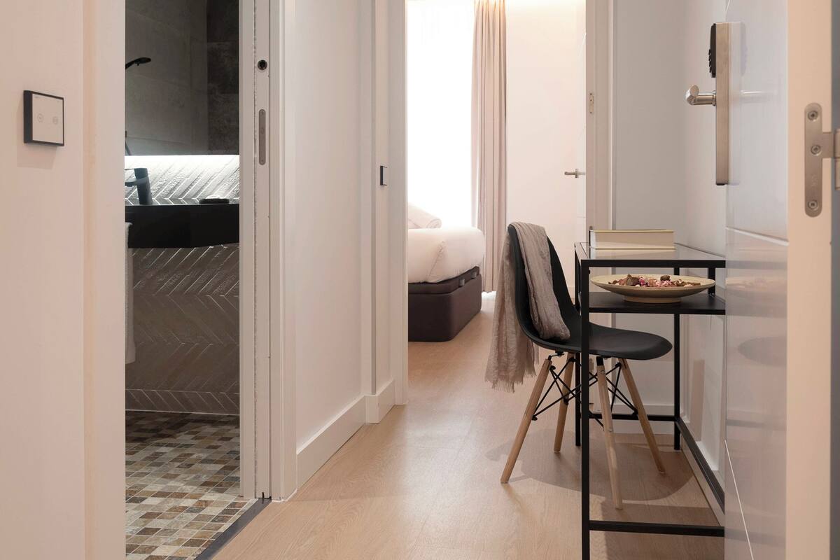 Een hotelkamer met een bed, een klein tafeltje met een schaal, een stoel en een deur naar een andere kamer.