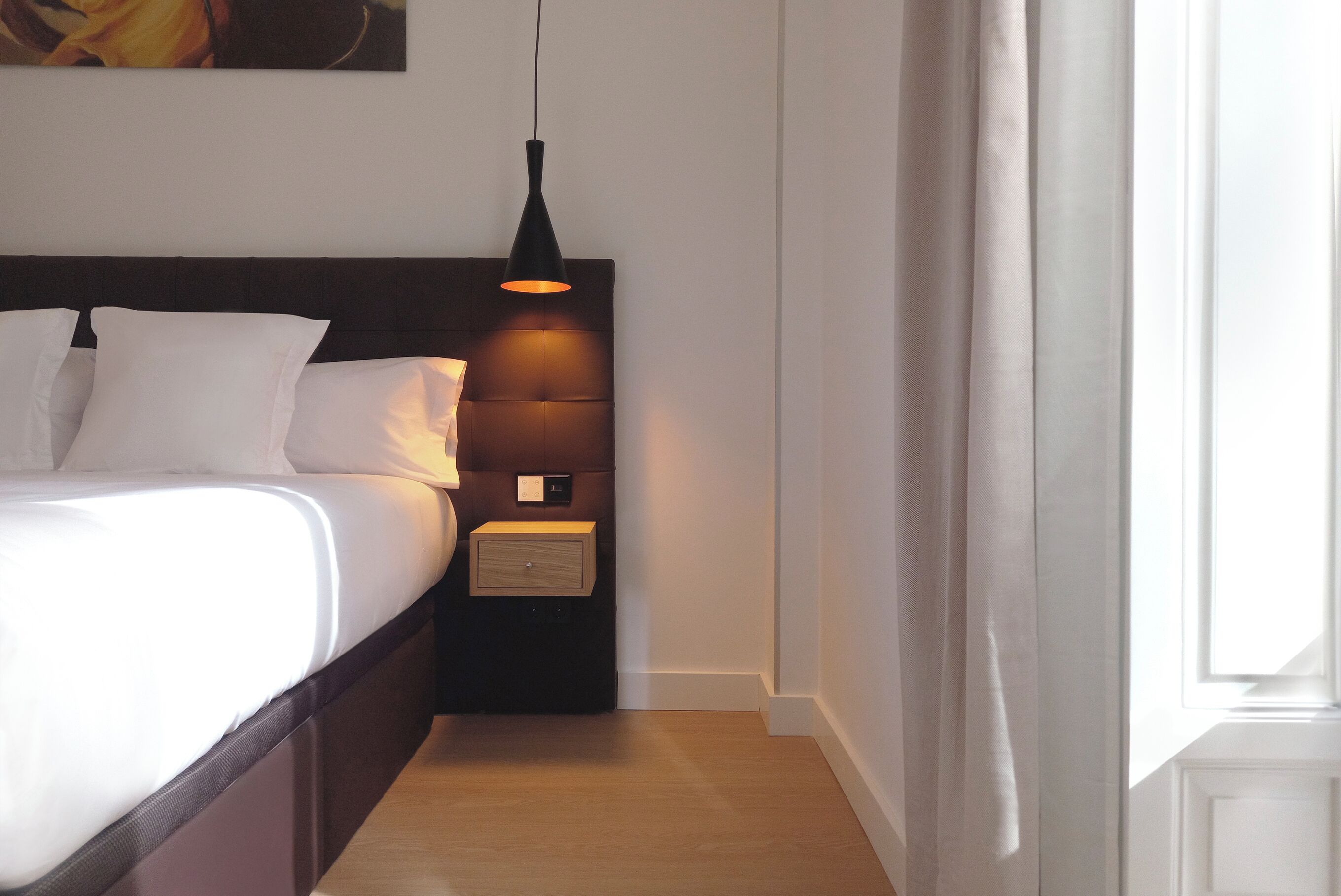 A neatly made bed with white linens, a bedside table with a lamp, and a wall-mounted artwork.