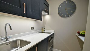 A modern kitchen with dark cabinetry, a white countertop, a stainless steel sink, a built-in oven, and a wall clock. - The Milan Suite at Moat House (Lancashire)