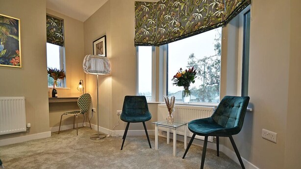 A cozy room with two teal chairs, a small table with a vase, and a window with a view of trees. - The Milan Suite at Moat House (Lancashire)