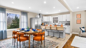 A modern kitchen with white cabinets, a stainless steel refrigerator, and a dining area with orange chairs and a wooden table. - 5bd Vibrant Haven: Basketball - 10mins to Airport (Charlotte)