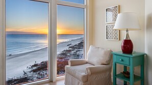 A room with a view of a beach at sunset, a white armchair, a teal side table with a red lamp, and two framed pictures on the wall. - Sunset Views, Gulf Front at Jade East Towers (Destin)