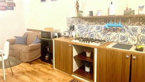 Une cuisine compacte équipée d’un four à micro-ondes, d’une bouilloire et d’une cuisinière à gaz. On y trouve un meuble en bois avec un évier intégré et une petite étagère où sont disposés des objets décoratifs. Un canapé marron et une chaise blanche se trouvent dans l’espace salon.