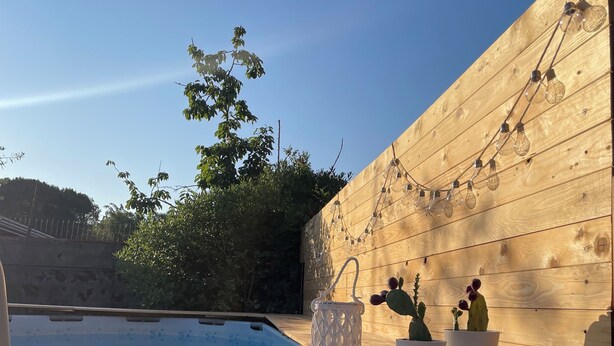 Une terrasse en bois avec une piscine, des plantes en pot et des guirlandes lumineuses.