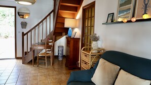 Una scala conduce a un secondo piano, una scrivania di legno con una sedia, una lampada blu e una poltrona in vimini.