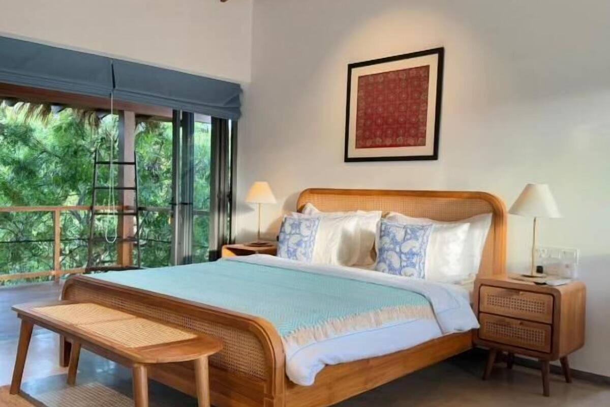 Een ruime slaapkamer met een groot bed, houten hoofdbord en uitzicht op groen door grote ramen.