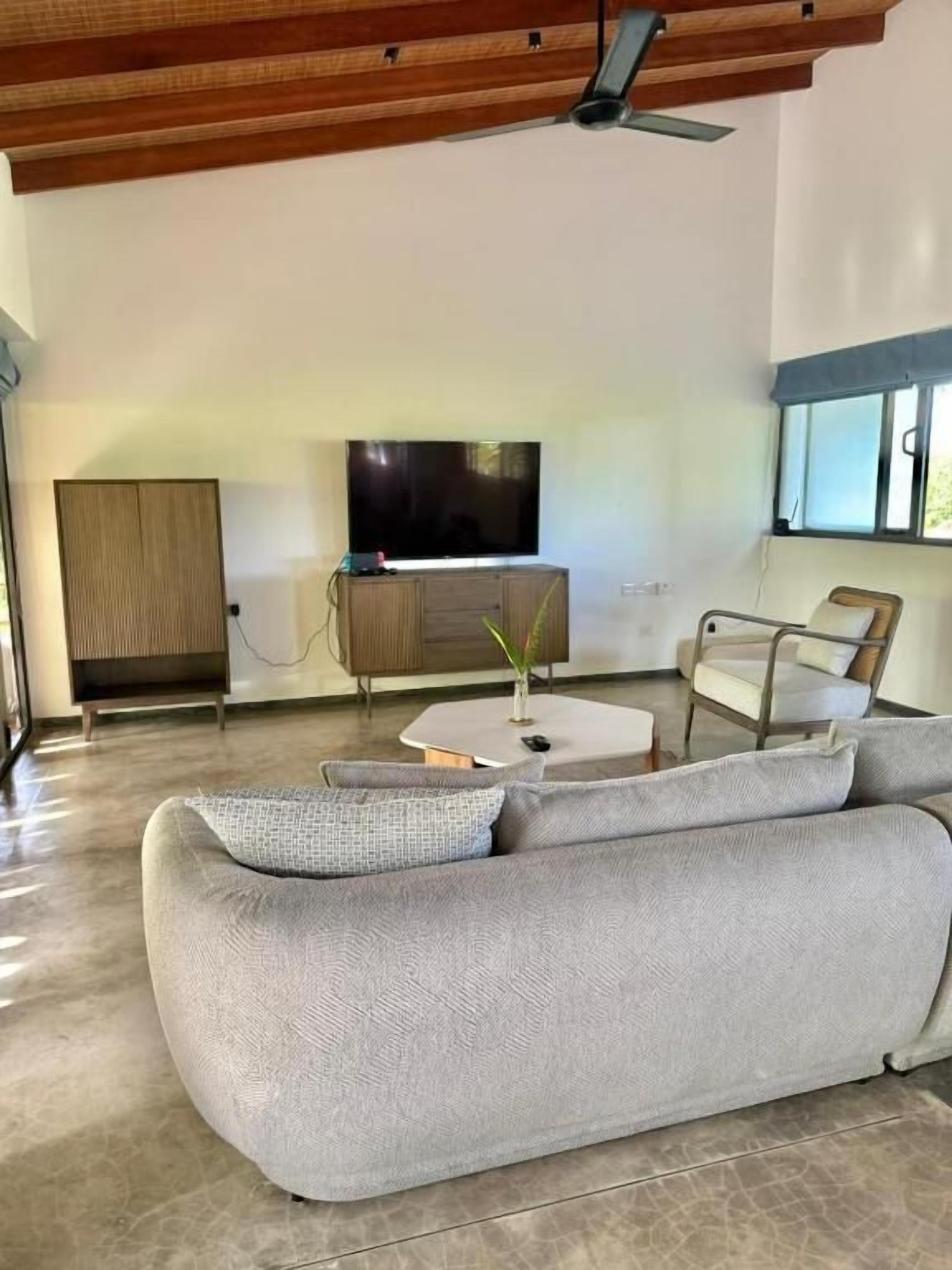 A modern living room with a grey sofa, a flat-screen TV, a wooden cabinet, and a window with blinds.