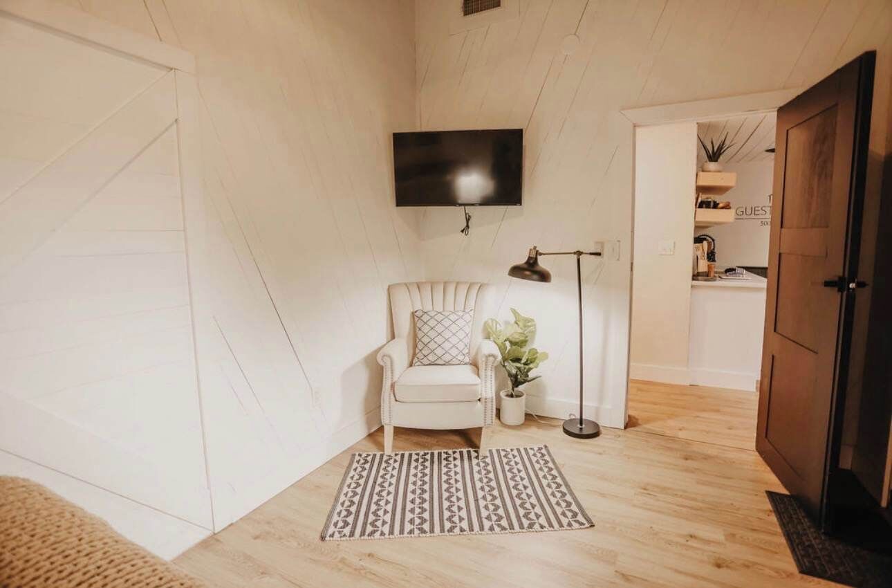 A room with a white chair, a flat-screen TV mounted on the wall, a floor lamp, and a patterned rug.
