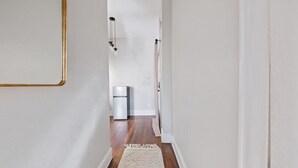 Un pasillo estrecho con suelo de madera, paredes blancas y una alfombra blanca. Se ve un refrigerador en la distancia.
