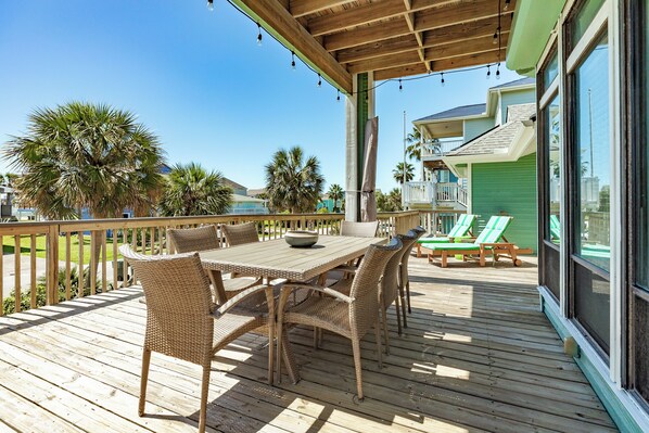 A wooden deck with a table and chairs, a railing, and a view of a green building with a balcony.