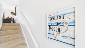 A staircase with beige carpeted steps and a white handrail, leading to a room with a wall-mounted lamp and a framed sign that reads 'see you at the beach'. - The Beachhouse - 2 Bed Townhouse - Saundersfoot (Saundersfoot)