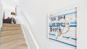 A staircase with beige carpeted steps and a white handrail, leading to a room with a wall-mounted lamp and a framed sign that reads 'see you at the beach'. - The Beachhouse - 2 Bed Townhouse - Saundersfoot (Saundersfoot)