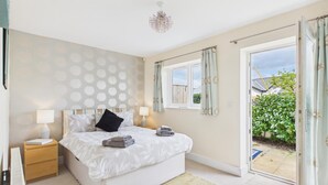 A bedroom with a bed, bedside tables, a chandelier, a window with curtains, and a view of a garden. - The Beachhouse - 2 Bed Townhouse - Saundersfoot (Saundersfoot)