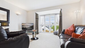 A living room with a TV, leather sofas, a glass coffee table, and a large window with curtains. - The Beachhouse - 2 Bed Townhouse - Saundersfoot (Saundersfoot)